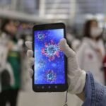 Gloved hand holding a smartphone displaying a 3D coronavirus illustration in a crowded public space.